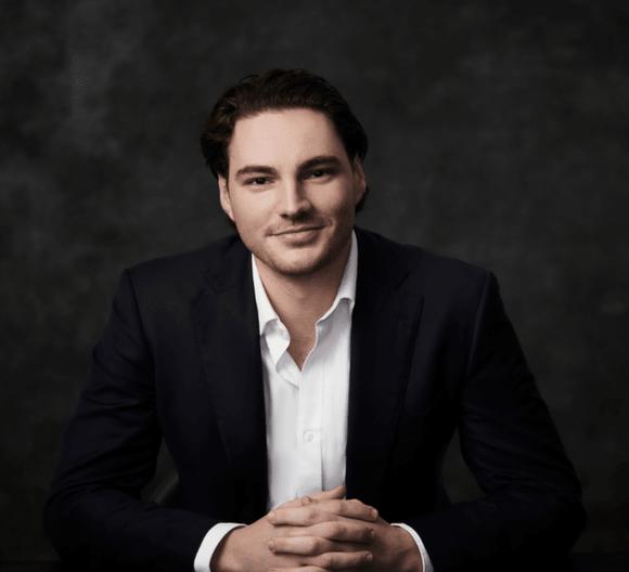 Professional portrait of a man in a dark suit sitting with hands clasped, against a textured background.
