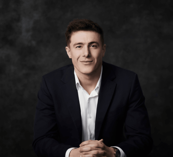 Confident young man in a business suit, sitting with hands clasped in front of a dark background.